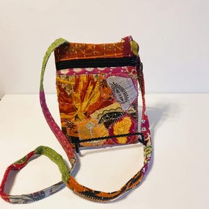 K&ouml;nnte beinhalten: Eine Patchwork-Umh&auml;ngetasche mit farbenfrohem Design. Die Tasche hat eine Mischung aus orangefarbenen, roten, gelben und rosa Stoffen, mit einem schwarzen Rei&szlig;verschluss und Riemen. Der Riemen ist ca. 102 cm lang.