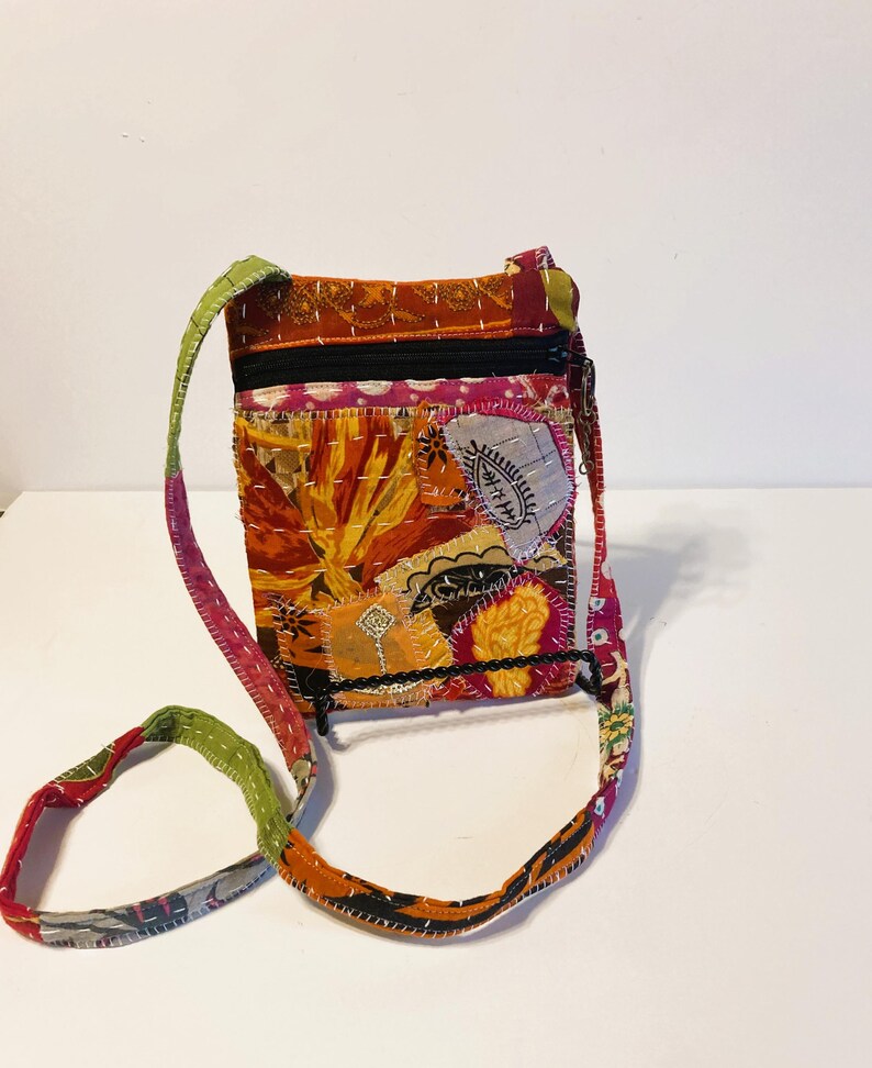 K&ouml;nnte beinhalten: Eine Patchwork-Umh&auml;ngetasche mit farbenfrohem Design. Die Tasche hat eine Mischung aus orangefarbenen, roten, gelben und rosa Stoffen, mit einem schwarzen Rei&szlig;verschluss und Riemen. Der Riemen ist ca. 102 cm lang.