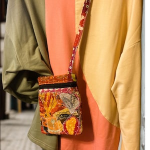 K&ouml;nnte beinhalten: Eine kleine, bunte Patchwork-Umh&auml;ngetasche mit floralem Riemen. Die Tasche hat eine Vielzahl von Stoffen in Orange-, Gelb- und Rott&ouml;nen mit einem schwarzen Rei&szlig;verschluss. Der Hintergrund ist ein farbblockiertes Oberteil in Olivgr&uuml;n, Koralle und Gelb.