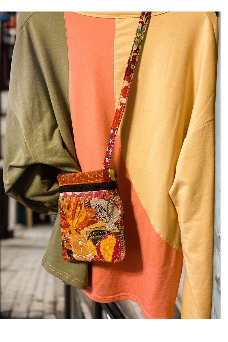 K&ouml;nnte beinhalten: Eine kleine, bunte Patchwork-Umh&auml;ngetasche mit floralem Riemen. Die Tasche hat eine Vielzahl von Stoffen in Orange-, Gelb- und Rott&ouml;nen mit einem schwarzen Rei&szlig;verschluss. Der Hintergrund ist ein farbblockiertes Oberteil in Olivgr&uuml;n, Koralle und Gelb.