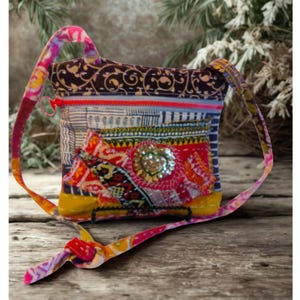May include: A colorful patchwork crossbody bag with a patterned strap. The bag features a mix of fabrics in various colors, including red, yellow, blue, and brown. A large sequined embellishment is centered on the front. The bag has a zipper closure.