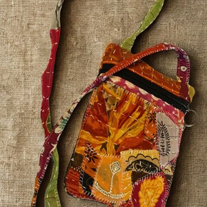 K&ouml;nnte beinhalten: Eine Patchwork-Umh&auml;ngetasche mit einem lebendigen Blumenmuster in Orange-, Gelb- und Rott&ouml;nen. Die Tasche hat einen schwarzen Rei&szlig;verschluss, einen kleinen Griff und einen langen Riemen mit einer Mischung aus bunten Mustern. Die Tasche ist etwa 18 cm breit und 23 cm hoch.