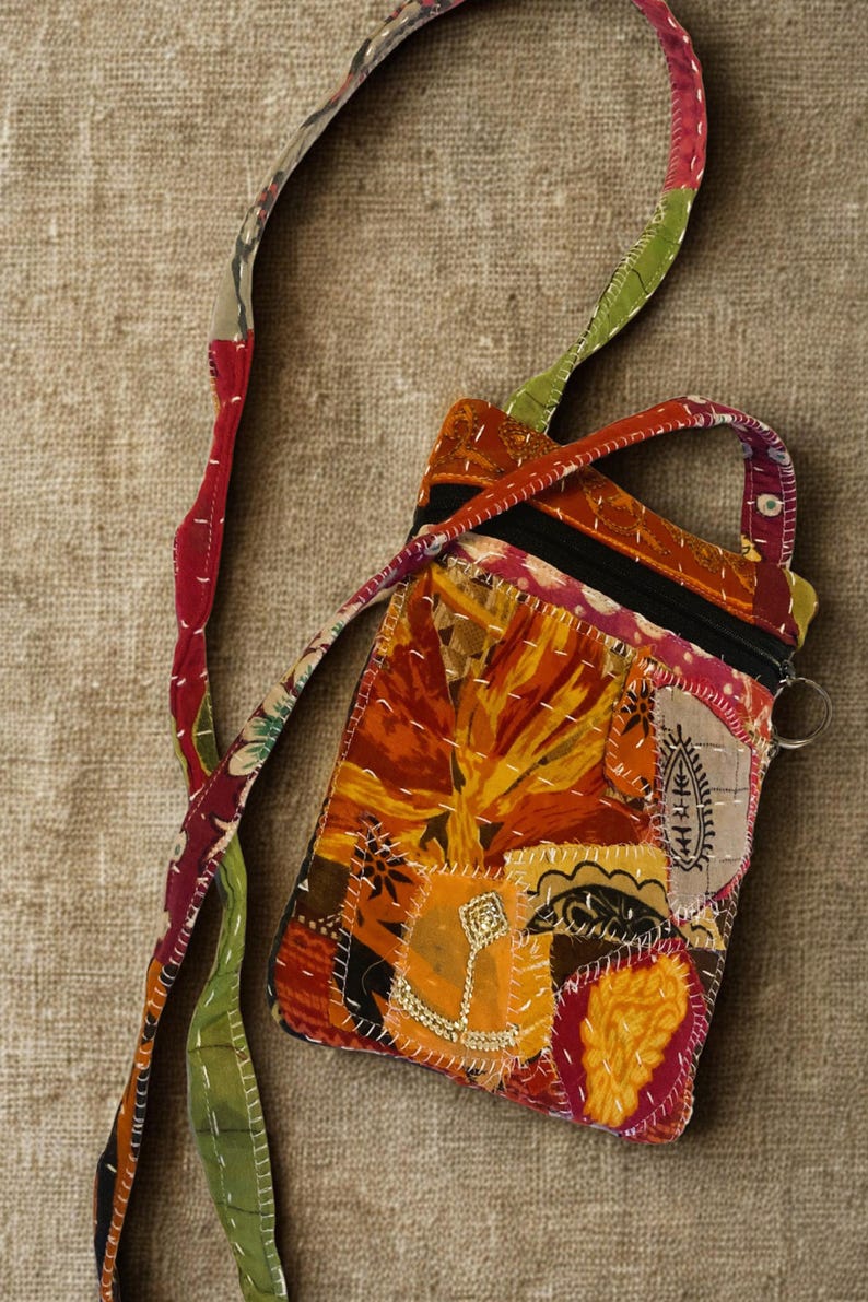 K&ouml;nnte beinhalten: Eine Patchwork-Umh&auml;ngetasche mit einem lebendigen Blumenmuster in Orange-, Gelb- und Rott&ouml;nen. Die Tasche hat einen schwarzen Rei&szlig;verschluss, einen kleinen Griff und einen langen Riemen mit einer Mischung aus bunten Mustern. Die Tasche ist etwa 18 cm breit und 23 cm hoch.