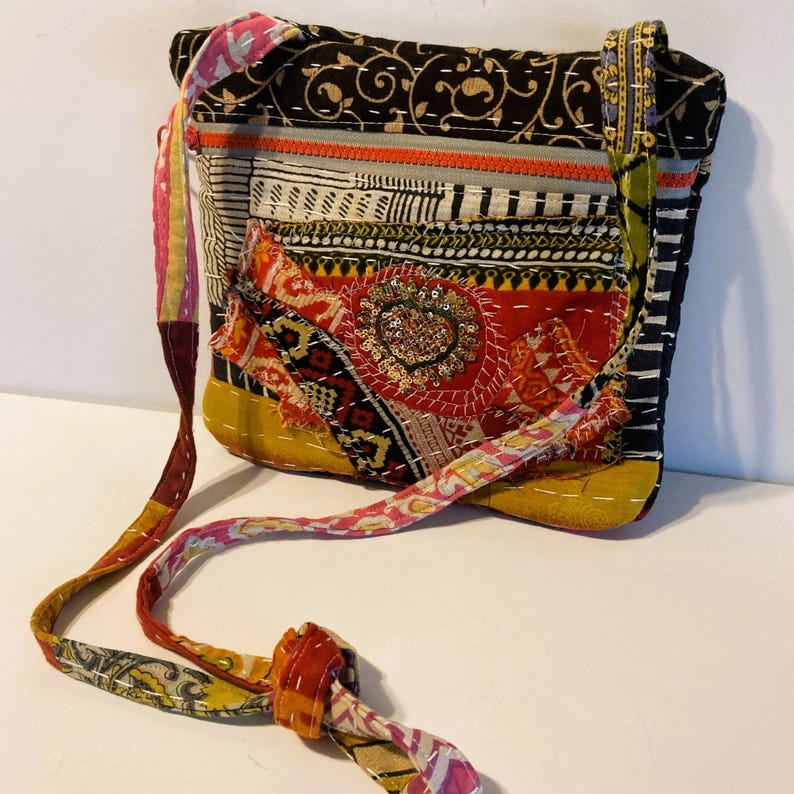 May include: A patchwork crossbody bag with a long strap. The bag features a variety of colorful fabric patterns, including floral, geometric, and striped designs. The bag's main colors are red, yellow, black, and pink, with decorative stitching throughout.