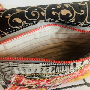 May include: Open view of a colorful, patterned bag with a red zipper. The bag's interior is visible, revealing a striped lining. The exterior features a mix of black, gold, red, and white patterns, suggesting a bohemian or ethnic design.