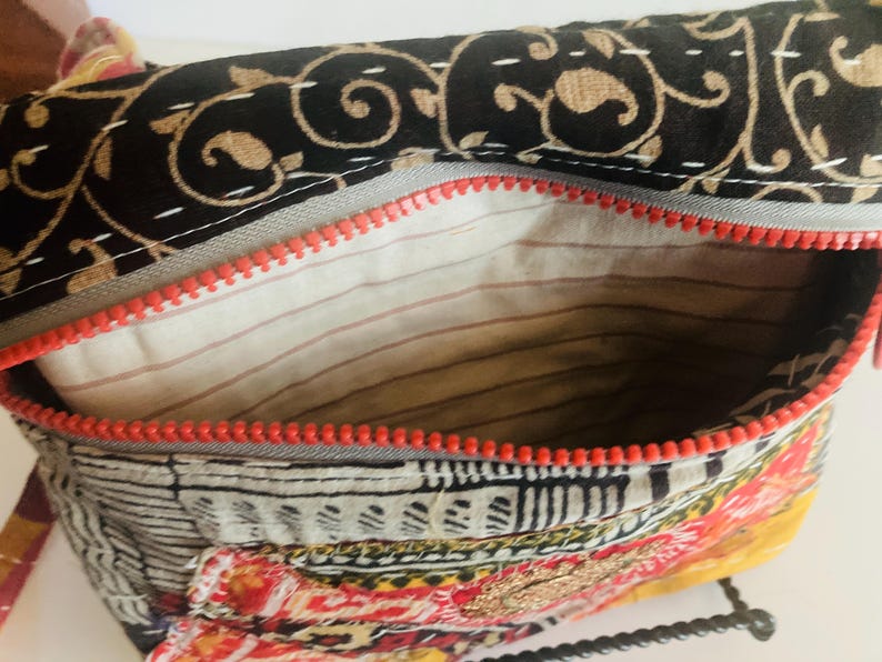 May include: Open view of a colorful, patterned bag with a red zipper. The bag's interior is visible, revealing a striped lining. The exterior features a mix of black, gold, red, and white patterns, suggesting a bohemian or ethnic design.