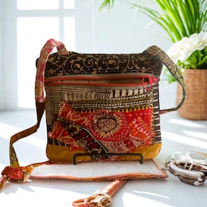 May include: A patchwork crossbody bag with a red zipper and strap. The bag features a mix of patterned fabrics in brown, red, and yellow, with a decorative heart-shaped embellishment. The bag has a long strap for over-the-shoulder wear.
