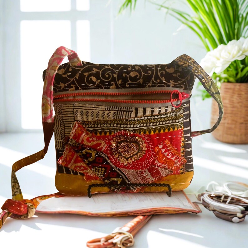 May include: A patchwork crossbody bag with a red zipper and strap. The bag features a mix of patterned fabrics in brown, red, and yellow, with a decorative heart-shaped embellishment. The bag has a long strap for over-the-shoulder wear.