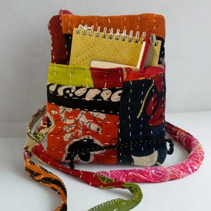 K&ouml;nnte beinhalten: Eine Patchwork-Umh&auml;ngetasche mit farbenfrohem, gestepptem Design. Die Tasche hat eine Vielzahl von Stoffen in Orange-, Rot-, Gr&uuml;n- und Blaut&ouml;nen mit wei&szlig;en N&auml;hten. Ein Spiralnotizbuch und ein Stift sind sichtbar. Der Riemen ist ca. 100 cm lang.
