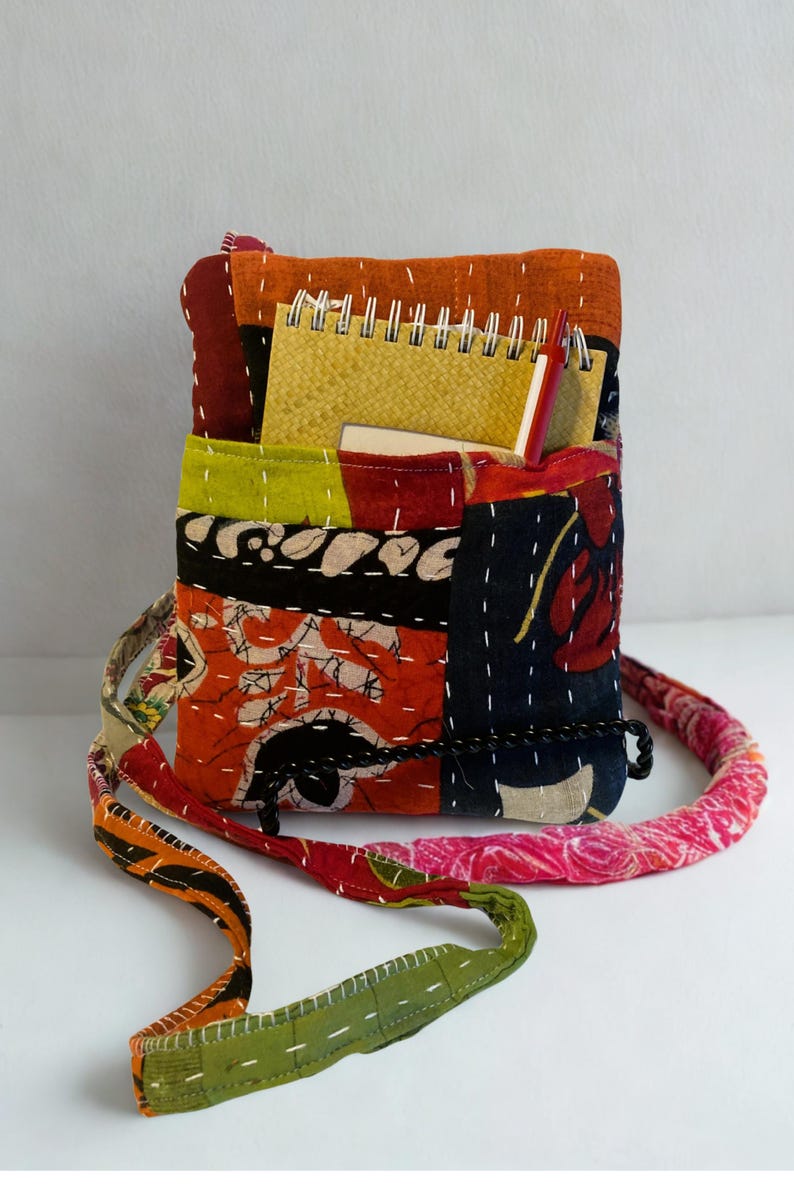K&ouml;nnte beinhalten: Eine Patchwork-Umh&auml;ngetasche mit farbenfrohem, gestepptem Design. Die Tasche hat eine Vielzahl von Stoffen in Orange-, Rot-, Gr&uuml;n- und Blaut&ouml;nen mit wei&szlig;en N&auml;hten. Ein Spiralnotizbuch und ein Stift sind sichtbar. Der Riemen ist ca. 100 cm lang.