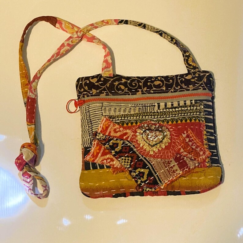 May include: A patchwork fabric bag with a patterned strap and a decorative tassel. The bag features a variety of colorful fabrics, including floral and geometric prints, with a sequined heart embellishment. The bag has a zipper closure.