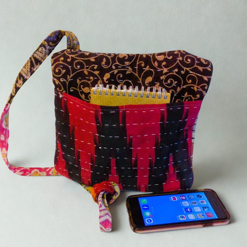 May include: A handmade, colorful shoulder bag with a red and black patterned front, a brown floral patterned top, and a patterned strap. A small yellow notepad is visible inside the bag's pocket. A smartphone rests in front of the bag.