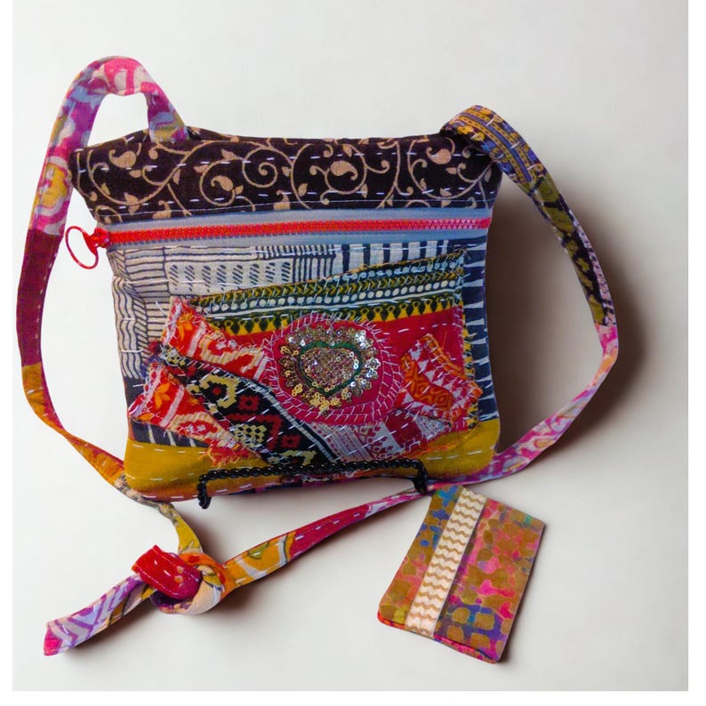 May include: A colorful patchwork crossbody bag with a patterned strap and a small matching tag. The bag features a variety of fabrics in red, yellow, blue, and brown, with a sequined heart-shaped embellishment. The bag has a zipper closure and a knotted strap.