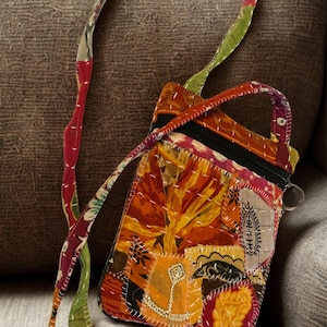 K&ouml;nnte beinhalten: Eine Patchwork-Umh&auml;ngetasche mit floralem Design in Orange-, Gelb- und Rott&ouml;nen. Die Tasche hat einen schwarzen Rei&szlig;verschluss und einen langen Riemen mit einer Mischung aus Mustern und Farben. Die Tasche ist ca. 15 cm breit und 20 cm hoch.