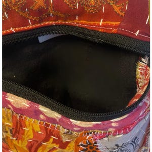 K&ouml;nnte beinhalten: Eine farbenfrohe, offene Handtasche mit schwarzem Rei&szlig;verschluss und Innenraum. Die Tasche hat ein Patchwork-Design mit Blumen- und geometrischen Mustern in Rot-, Orange- und Rosat&ouml;nen. Wei&szlig;e N&auml;hte akzentuieren den Stoff.