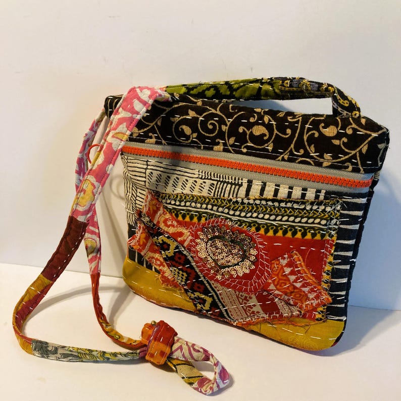 May include: A patchwork shoulder bag with a variety of patterns and colors. The bag features a black background with gold floral designs, a yellow base, and a red and orange patch with sequins. The strap is multi-colored.