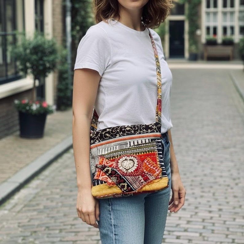 May include: A colorful crossbody bag with a patchwork design. The bag features a mix of patterns, including floral and geometric motifs, in shades of red, black, and yellow. The strap is also patterned. The bag is worn over the shoulder.
