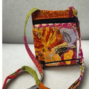 K&ouml;nnte beinhalten: Eine Patchwork-Umh&auml;ngetasche mit farbenfrohem Design. Die Tasche hat eine Mischung aus orangefarbenen, gelben und rosa Stoffen mit einem schwarzen Rei&szlig;verschluss und Riemen. Der Riemen ist eine Mischung aus gr&uuml;nen, roten und orangefarbenen Stoffen.