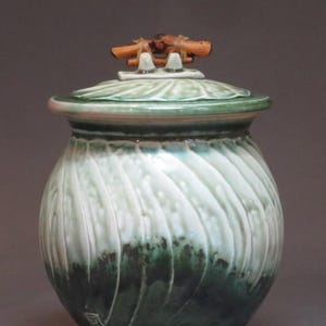 May include: A handcrafted ceramic jar with a lid. The jar is a swirl design with shades of green and white. The lid has a leaf design and a handle made of small wooden pieces. The jar is approximately 8 inches tall.