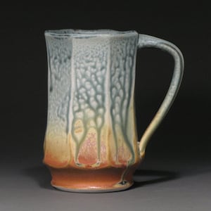May include: A tall, handmade ceramic mug with a unique, textured glaze. The mug features a vertical, faceted design with a handle. The colors transition from a light blue at the top to a warm orange-brown at the base, with green and pink accents.