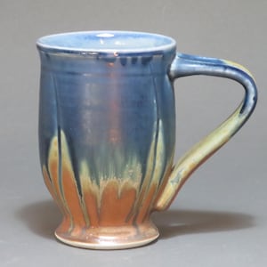 May include: A handmade ceramic mug with a blue exterior and a contrasting handle. The mug features a unique design with orange and yellow accents, adding visual interest. The mug is designed for hot beverages.
