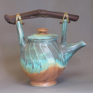 May include: A handcrafted ceramic teapot with a wooden handle. The teapot features a teal, brown, and gray color scheme with a textured surface. The lid and handle are brown, and the handle is attached with yellow ties. The teapot has a spout.