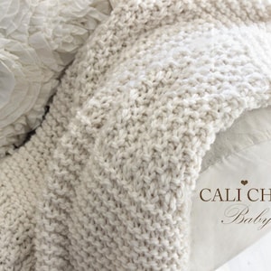 Puede incluir: Manta de punto grueso de color crema, sobre una superficie blanca. La manta tiene un aspecto texturizado y tejido. En el fondo se ve un cojín decorativo con tela con volantes. Las palabras "Cali Chic Baby" están impresas en la imagen.