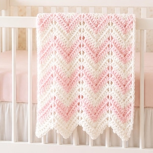 May include: A handmade crochet baby blanket featuring a repeating chevron pattern in alternating stripes of soft pastel pink and creamy white yarn. The textile has a textured, open-stitch design with a scalloped bottom edge draped over a white crib.