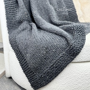 Pode incluir: Um cobertor de malha cinza escuro com uma aparência texturizada e manchas brancas. O cobertor é dobrado sobre uma superfície branca, mostrando sua malha grossa e aconchegante. O cobertor tem uma borda grossa e é rotulado como "Cali Chic Baby".
