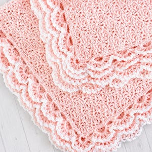 May include: A soft, light pink crocheted blanket featuring a textured, repeating shell stitch pattern throughout the center. The edges are finished with a decorative, scalloped white crochet border that creates a distinct, layered effect.