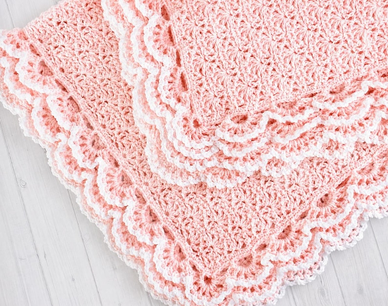 May include: A soft, light pink crocheted blanket featuring a textured, repeating shell stitch pattern throughout the center. The edges are finished with a decorative, scalloped white crochet border that creates a distinct, layered effect.