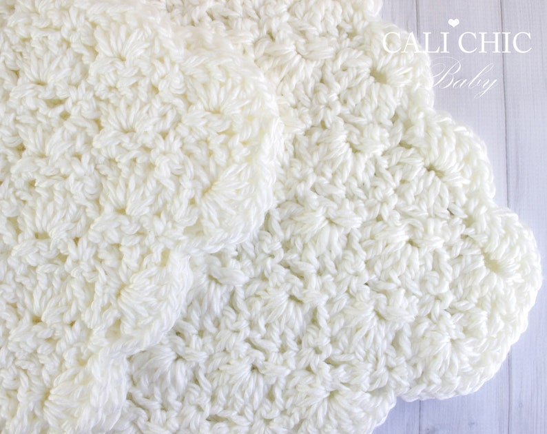 May include: A white crocheted baby blanket with a scalloped edge. The blanket is made with a delicate stitch pattern and is soft and cozy.