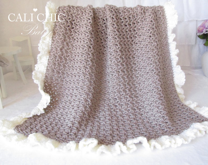 May include: A brown crocheted baby blanket with a white crocheted lace trim. The blanket is folded in half and has a "Cali Chic Baby" label in the top left corner.