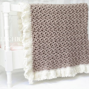 May include: A crocheted baby blanket in a light brown color with a white crocheted border and ruffle. The blanket is displayed on a white crib.