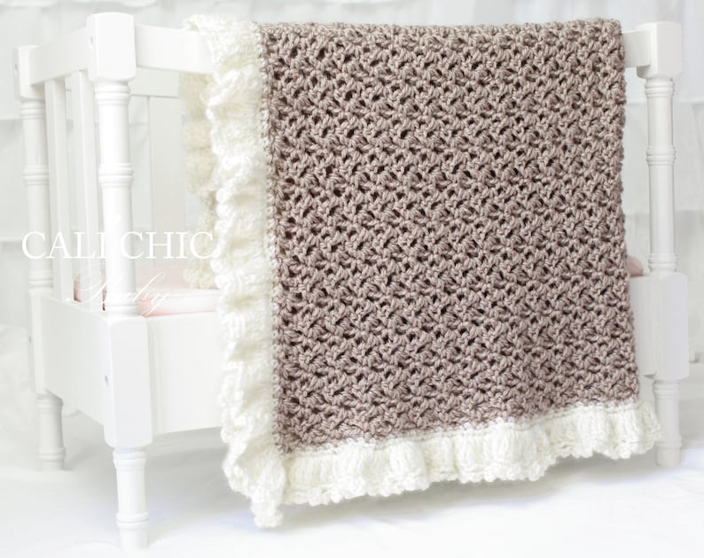 May include: A crocheted baby blanket in a light brown color with a white crocheted border and ruffle. The blanket is displayed on a white crib.