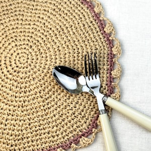 May include: A woven placemat with a scalloped edge in beige and mauve, paired with a fork and spoon. The silverware has cream-colored handles. The placemat is round and has a spiral design.