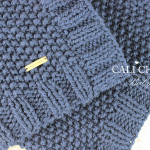 Op de afbeelding: Een donkerblauwe gebreide babydeken met een geweven label waarop "Cali Chic" staat. De deken heeft een textuurpatroon en is effen gekleurd.