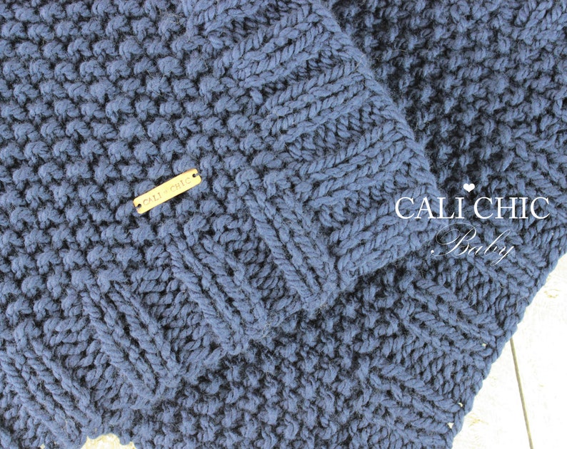 Op de afbeelding: Een donkerblauwe gebreide babydeken met een geweven label waarop "Cali Chic" staat. De deken heeft een textuurpatroon en is effen gekleurd.