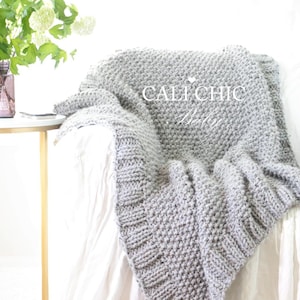 Puede incluir: Una manta gris tejida con un patrón de punto grueso y una orilla acanalada, sobre una superficie blanca. El texto "CALI CHIC Baby" es visible en la manta. Una pequeña mesa auxiliar con un jarrón de flores está en el fondo.