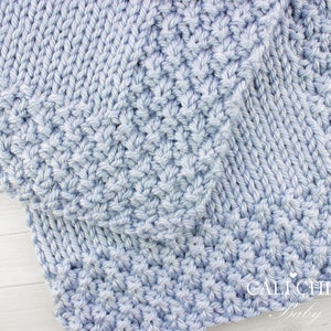 May include: Close-up of a light blue knitted baby blanket with a textured stitch pattern. The blanket is folded in half and the edge of the blanket is visible. The text "CALI CHIC Baby" is visible in the bottom right corner of the image.