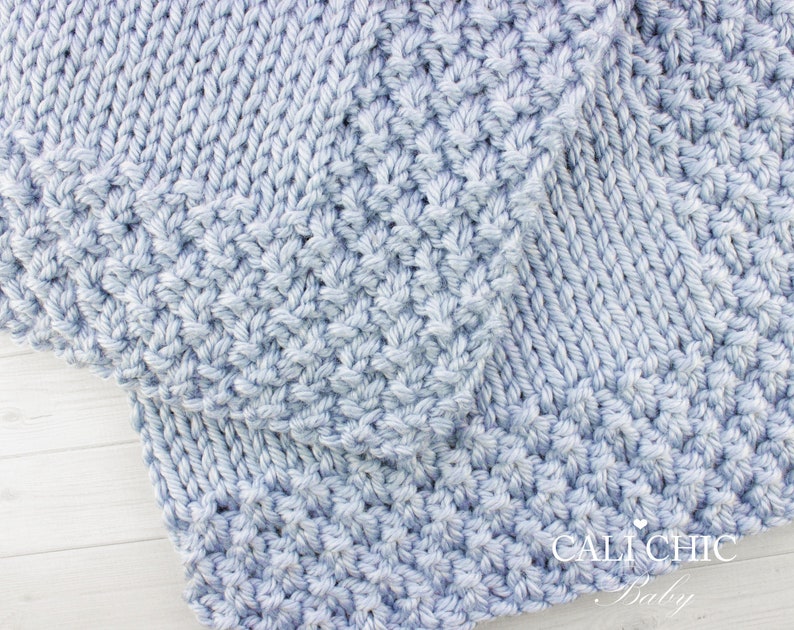 May include: Close-up of a light blue knitted baby blanket with a textured stitch pattern. The blanket is folded in half and the edge of the blanket is visible. The text "CALI CHIC Baby" is visible in the bottom right corner of the image.