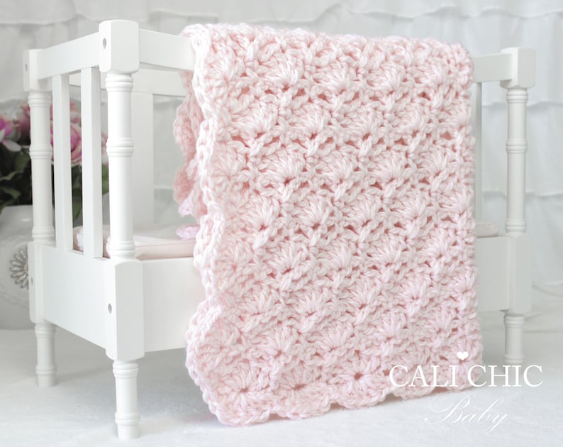 May include: A pink crocheted baby blanket with a scalloped edge, draped over a white crib.