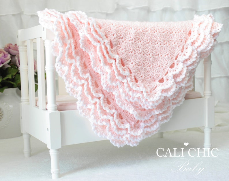 May include: A pink crocheted baby blanket with white trim and scalloped edges. The blanket is folded and displayed on a white crib. The text 'CALI CHIC Baby' is visible in the lower right corner of the image.