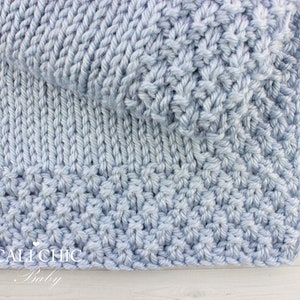 May include: A light blue knitted baby blanket with a textured stitch pattern. The blanket is folded in half and the bottom edge is visible. The text "CALI CHIC Baby" is visible in the bottom left corner.