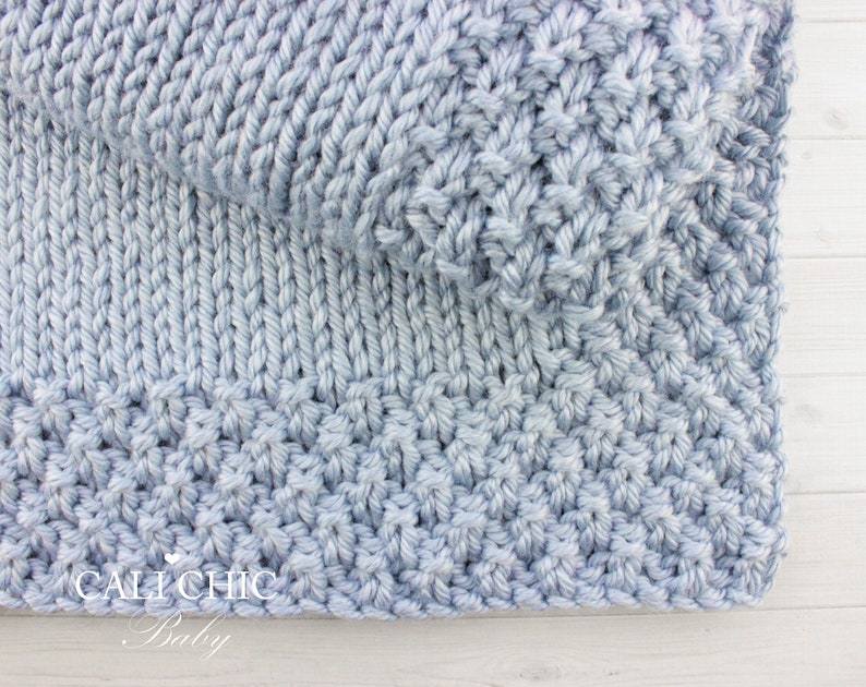 May include: A light blue knitted baby blanket with a textured stitch pattern. The blanket is folded in half and the bottom edge is visible. The text "CALI CHIC Baby" is visible in the bottom left corner.