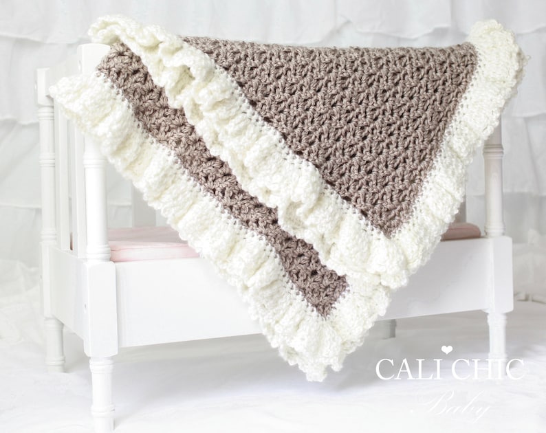 May include: A crocheted baby blanket with a white ruffle border and a brown center. The blanket is draped over a white crib with a pink mattress.