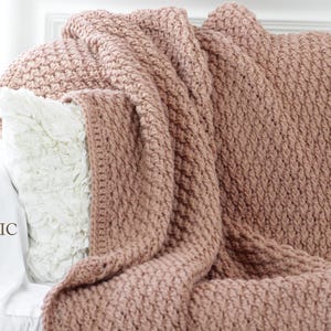 Pode incluir: Um cobertor de malha grossa cor de rosa empoeirado sobre roupa de cama e uma almofada brancas. O cobertor tem um padrão tecido texturizado. As palavras "Cali Chic Baby" estão impressas na roupa de cama.