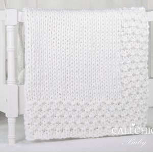 May include: White knitted baby blanket draped over a white crib. The blanket has a textured knit pattern and a small heart with the text "Cali Chic Baby" in the bottom right corner.