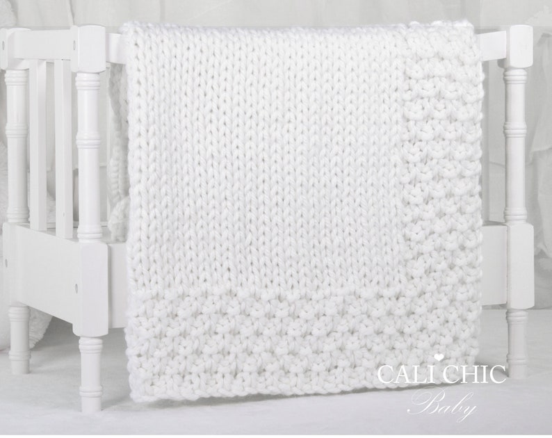 May include: White knitted baby blanket draped over a white crib. The blanket has a textured knit pattern and a small heart with the text "Cali Chic Baby" in the bottom right corner.