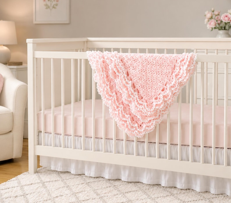 May include: A light pink crocheted baby blanket with a ruffled, scalloped edge draped over the side of a white wooden crib. The crib features a light pink mattress sheet and a white ruffled bed skirt, creating a soft and layered appearance.